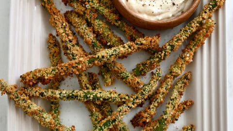 A top-down view of Baked Asparagus Fries on a white serving platter next to a small bowl containing roasted garlic aioli