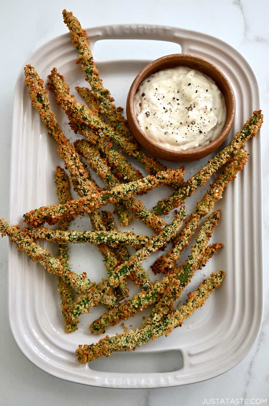 A top-down view of Baked Asparagus Fries on a white serving platter next to a small bowl containing roasted garlic aioli