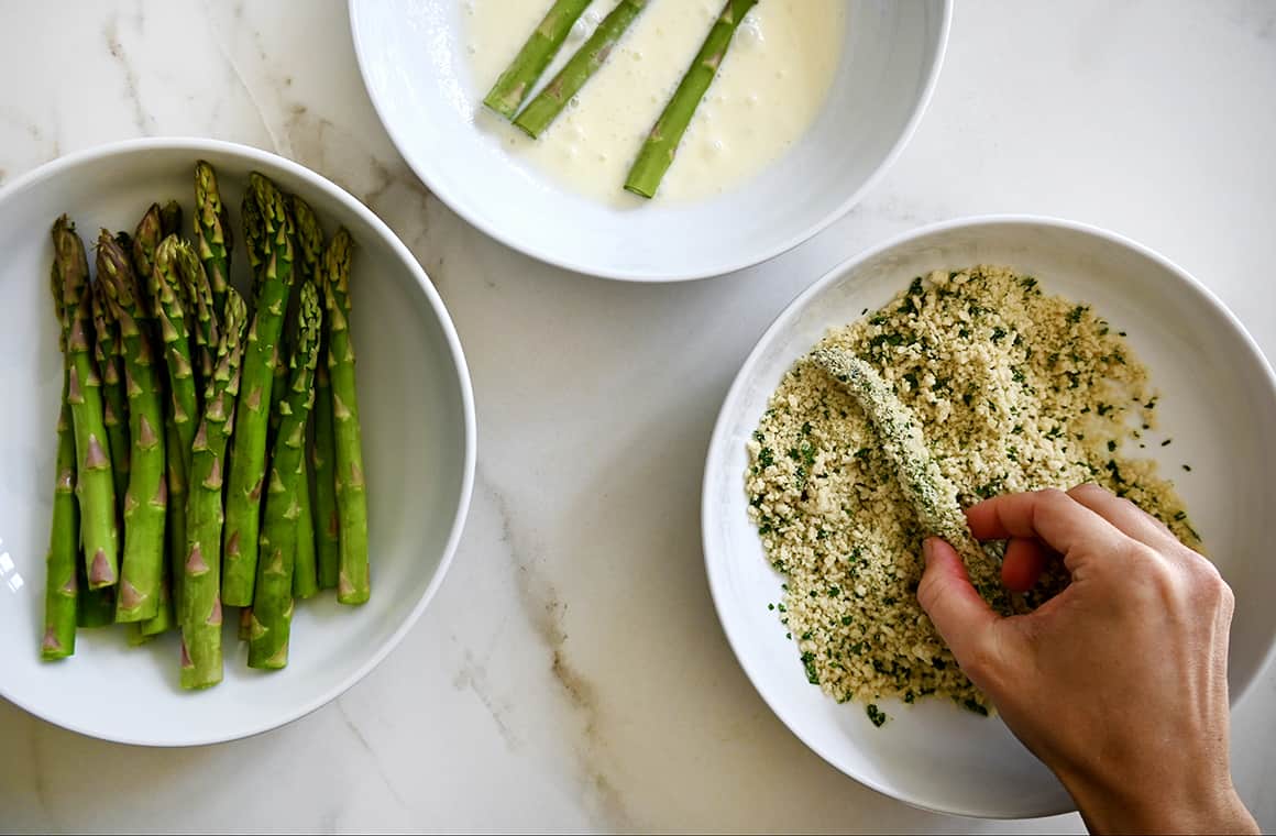 A top-down view of three bowls containing asparagus spears, an egg wash and breadcrumbs