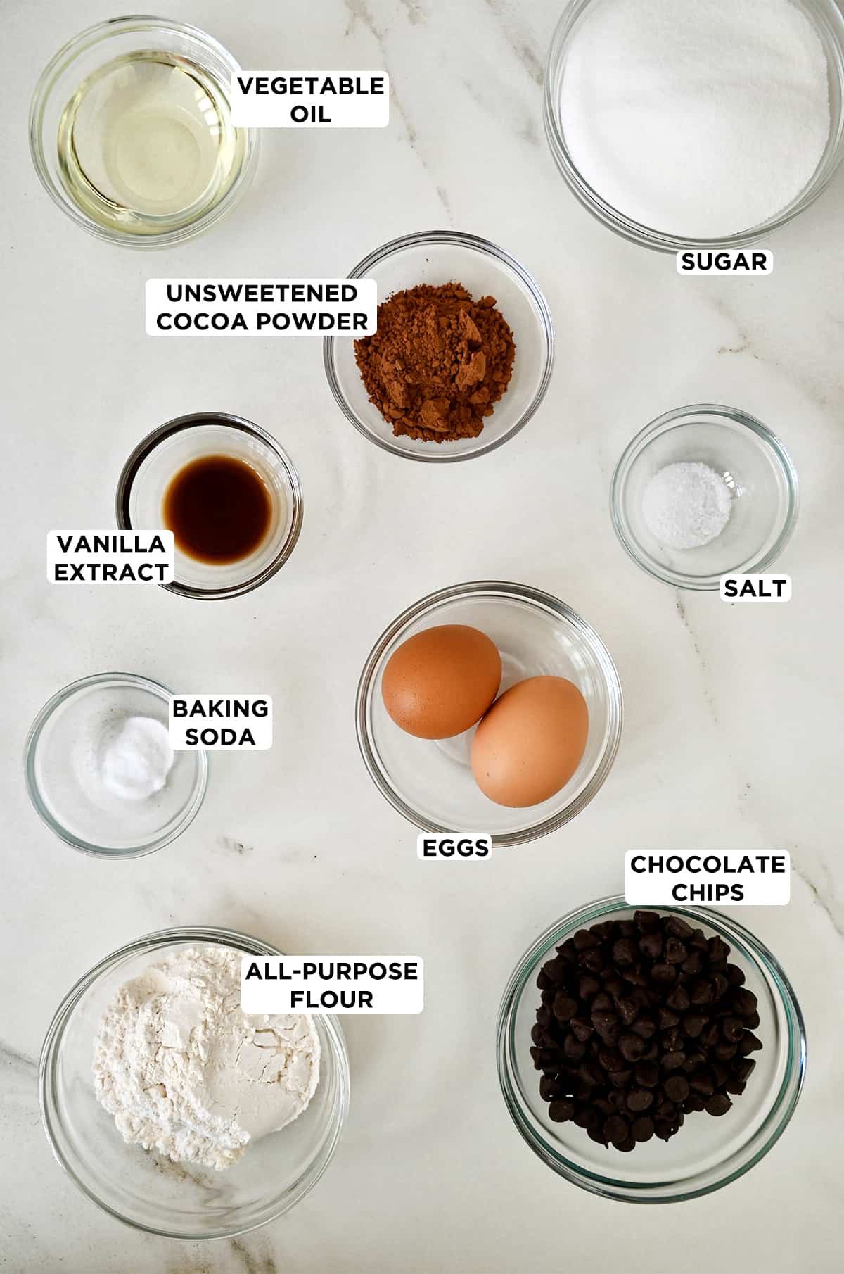 Glass bowls containing the ingredients to make brownie brittle, including sugar, salt, eggs, chocolate chips, all-purpose flour, baking soda, vanilla extract, unsweetened cocoa powder and vegetable oil.