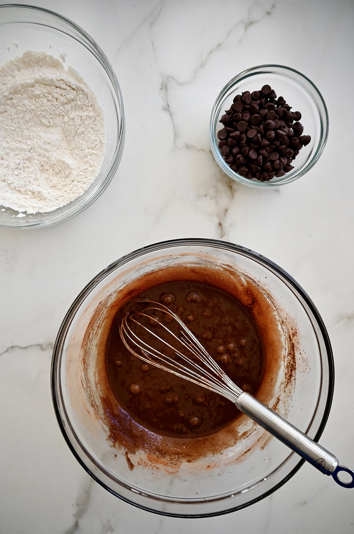 Three glass bowls containing flour, brownie batter with a whisk, and chocolate chips.