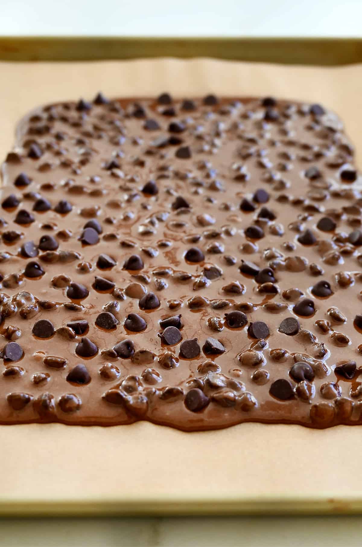 Brownie batter spread thinly atop parchment paper on a baking sheet.
