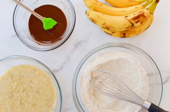 Bowl containing melted chocolate with spatula next to bunch of bananas, bowl with dry ingredients and whisk, and bowl with mashed bananas