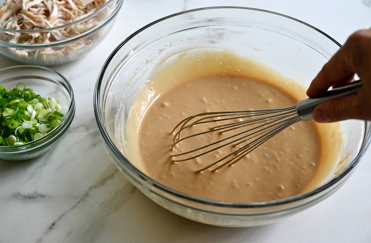 A hand holds a whisk into a clear bowl containing peanut sauce next to a small bowl with chopped scallions.