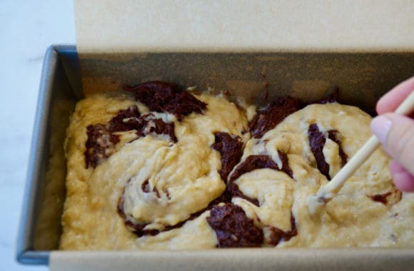 Hand with wooden dowel swirling together banana batter and the chocolate batter in bread pan