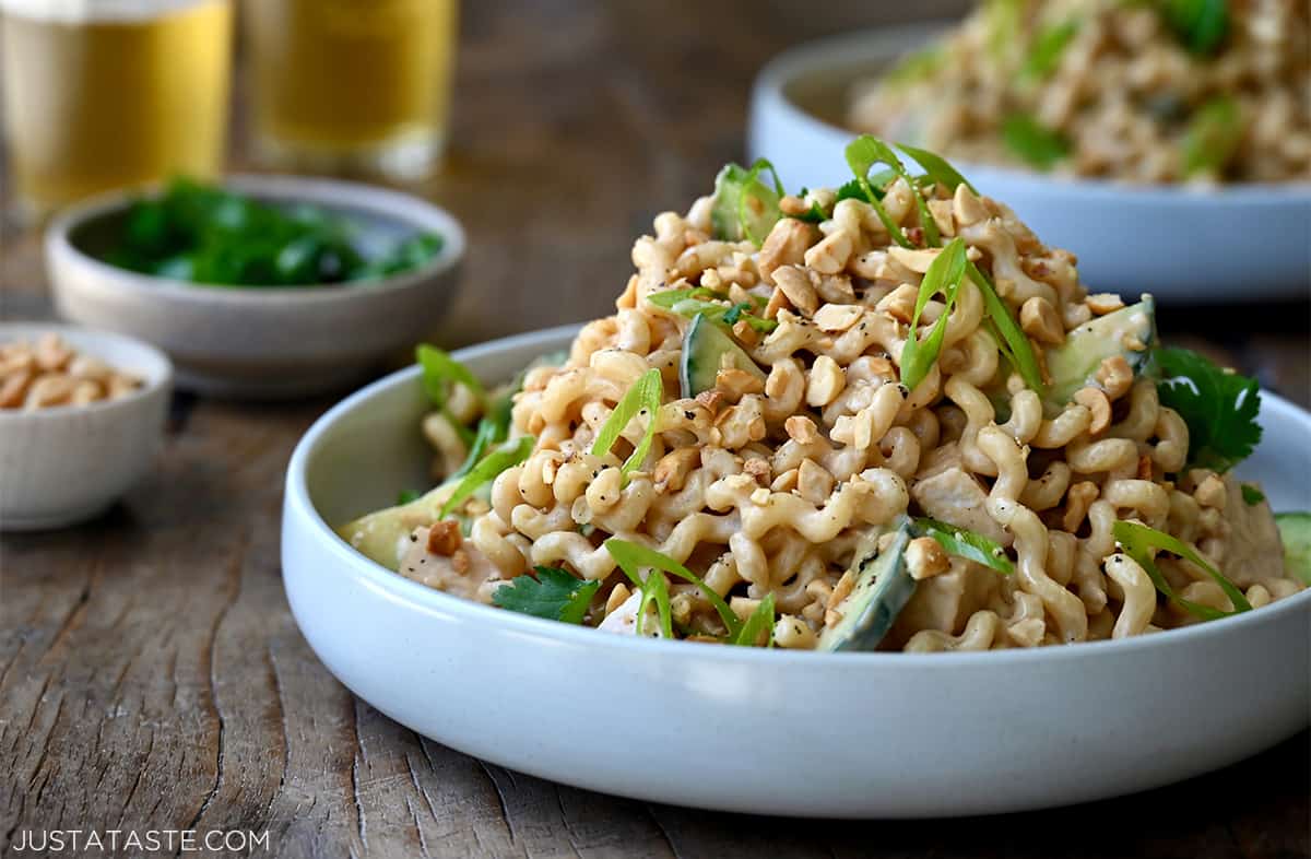 A white dinner plate piled high with Thai-inspired pasta salad with rotisserie chicken, sliced cucumbers and scallions.