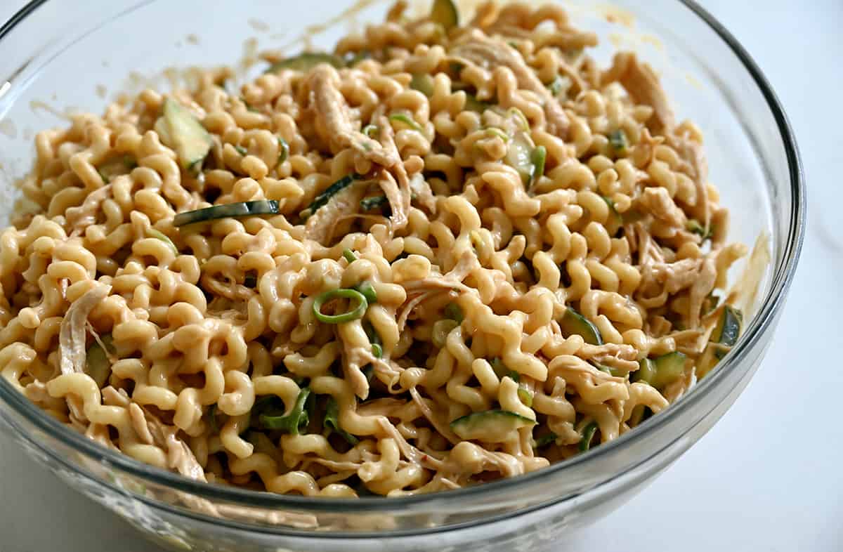 A large clear bowl containing spiral noodles and sliced scallions tossed in Thai peanut sauce.