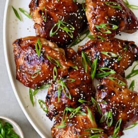 Honey garlic chicken thighs garnished with sesame seeds and sliced scallions on a white serving platter.
