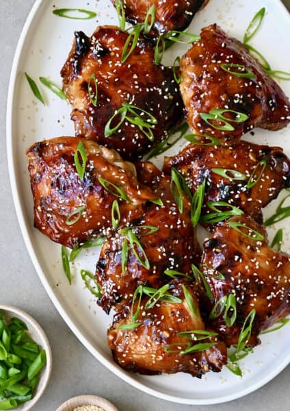 Honey garlic chicken thighs garnished with sesame seeds and sliced scallions on a white serving platter.