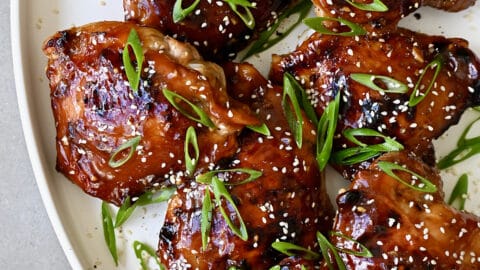 Honey garlic chicken thighs garnished with sesame seeds and sliced scallions on a white serving platter.