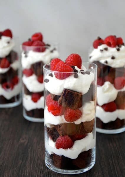 A dessert cup with layers of brownie chunks, whipped cream and fresh raspberries topped with mini chocolate chips.