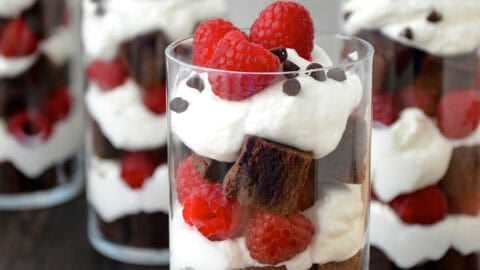 A dessert cup with layers of brownie chunks, whipped cream and fresh raspberries topped with mini chocolate chips.