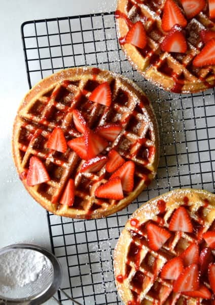 Three Belgian waffles topped with fresh strawberries, strawberry syrup and powdered sugar on a wire cooling rack.