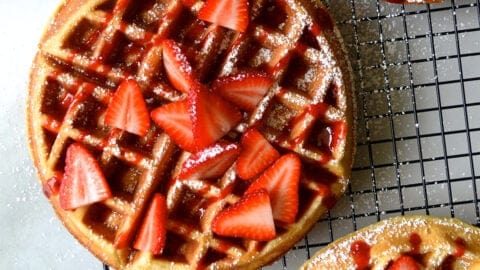 Three Belgian waffles topped with fresh strawberries, strawberry syrup and powdered sugar on a wire cooling rack.