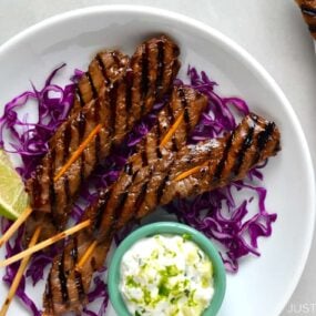 A white plate containing purple shredded cabbage and beef kabobs on skewers