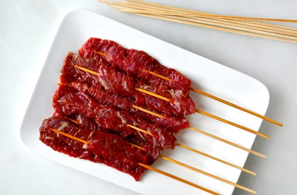 A white plate containing marinated beef kabobs on wooden skewers