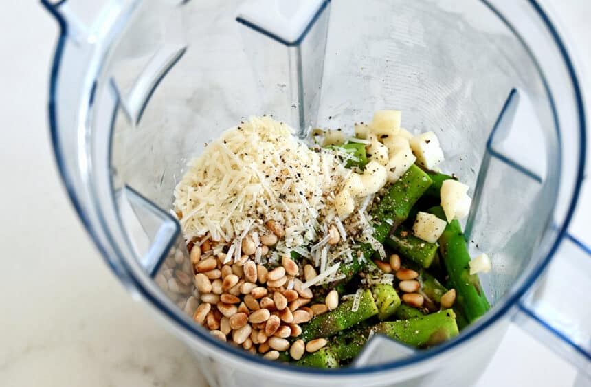A top-down view of a blender containing toasted pine nuts, shredded Parmesan cheese, garlic cloves, salt and pepper
