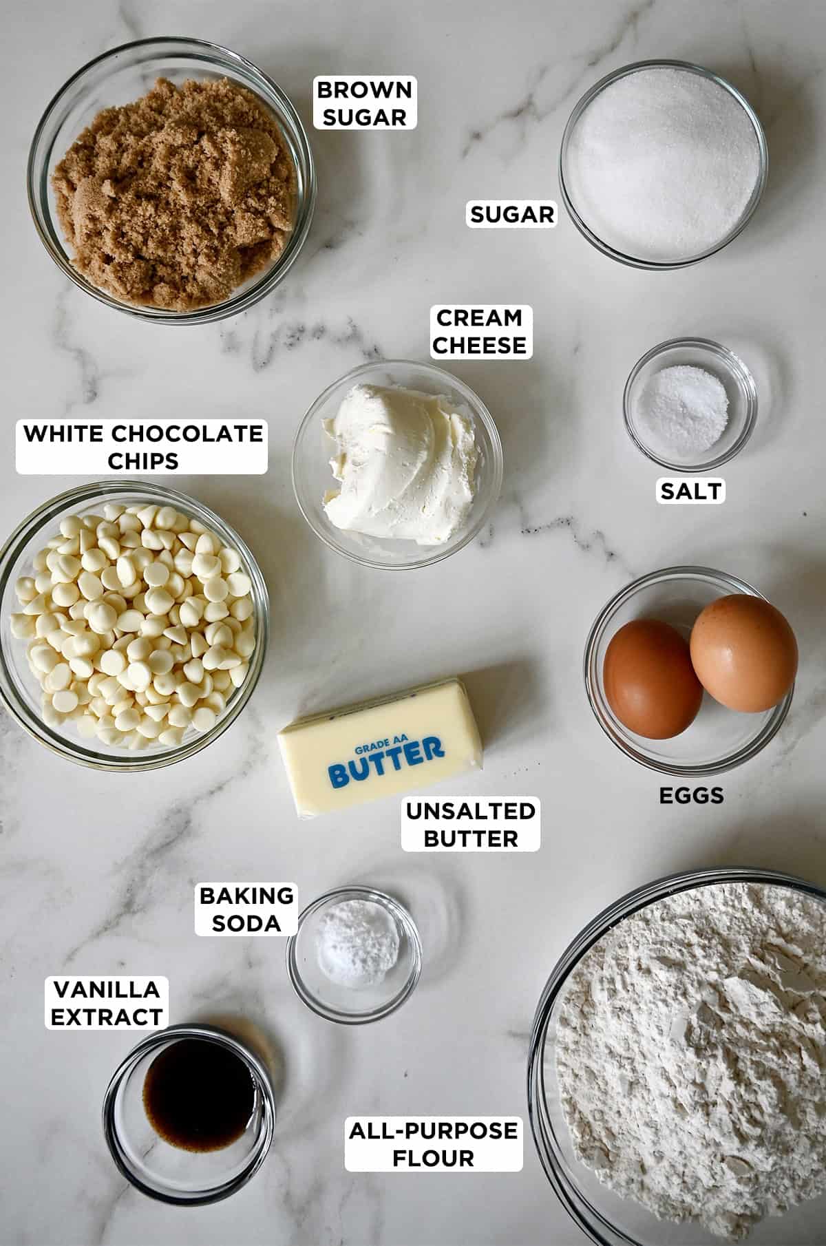 Ingredients to make white chocolate cream cheese cookies in various sizes of glass bowls: brown and granulated sugar, salt, cream cheese, two eggs, a stick of butter, white chocolate chips, baking soda, vanilla extract and flour.