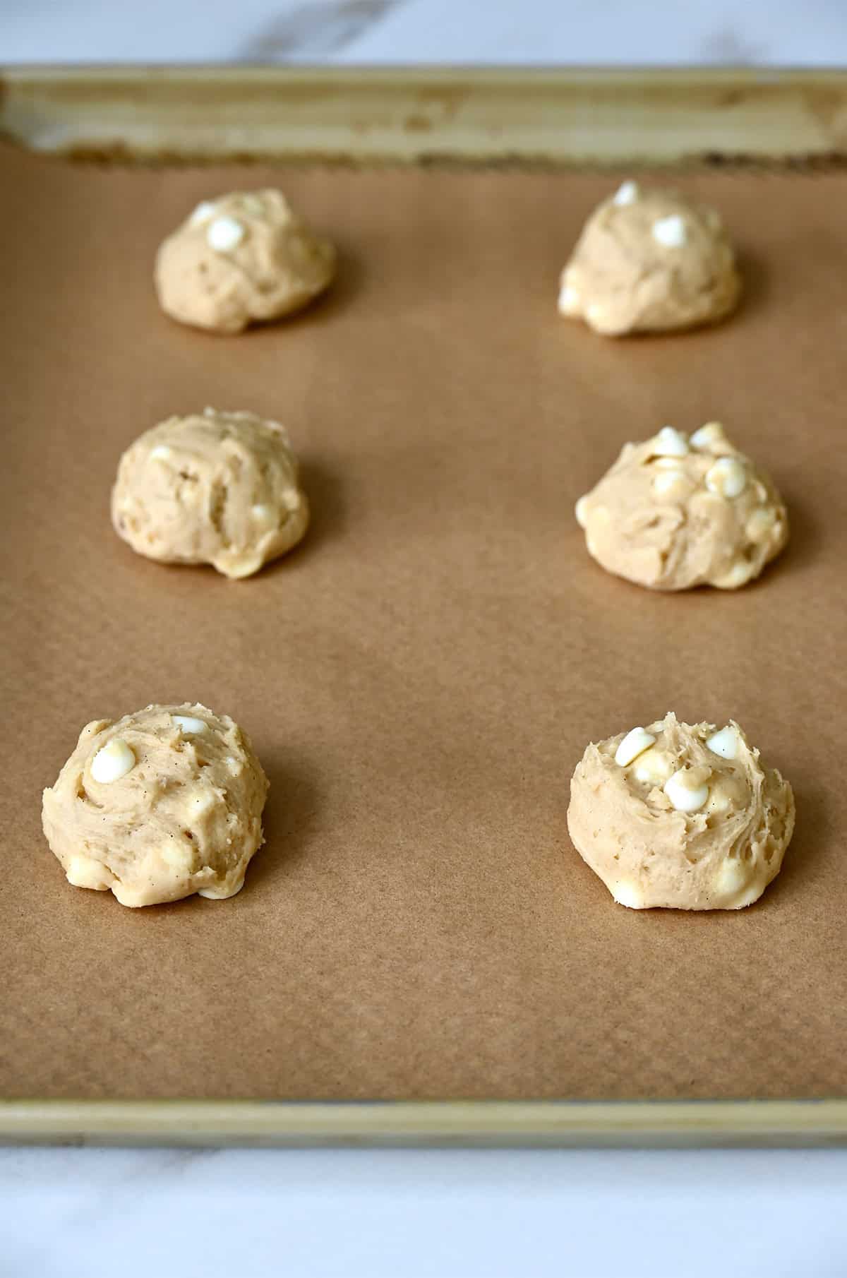 Mounds of cheesecake cookie dough studded with white chocolate chips on a parchment paper lined baking sheet.
