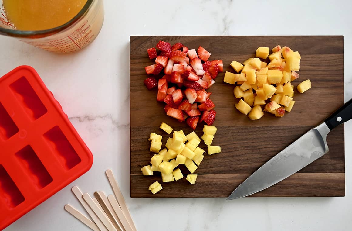 Chopped fruit on a cutting board with a sharp knife next to popsicle sticks and a popsicle mold