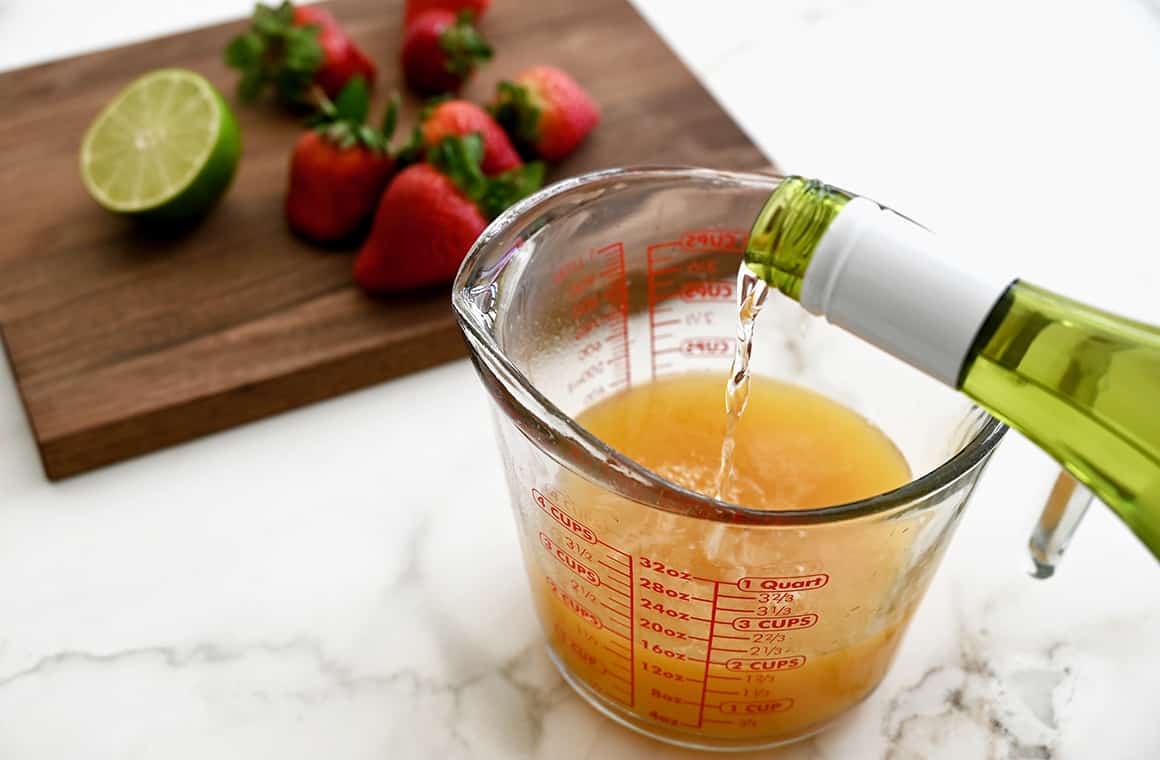 White wine being poured into a liquid measuring cup with fresh fruit on a cutting board in the background 