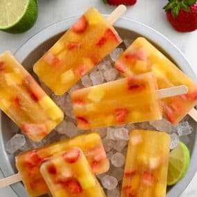 A top-down view of Peach Sangria Popsicles over ice in a pie plate next to fresh strawberries and a lime cut open in half