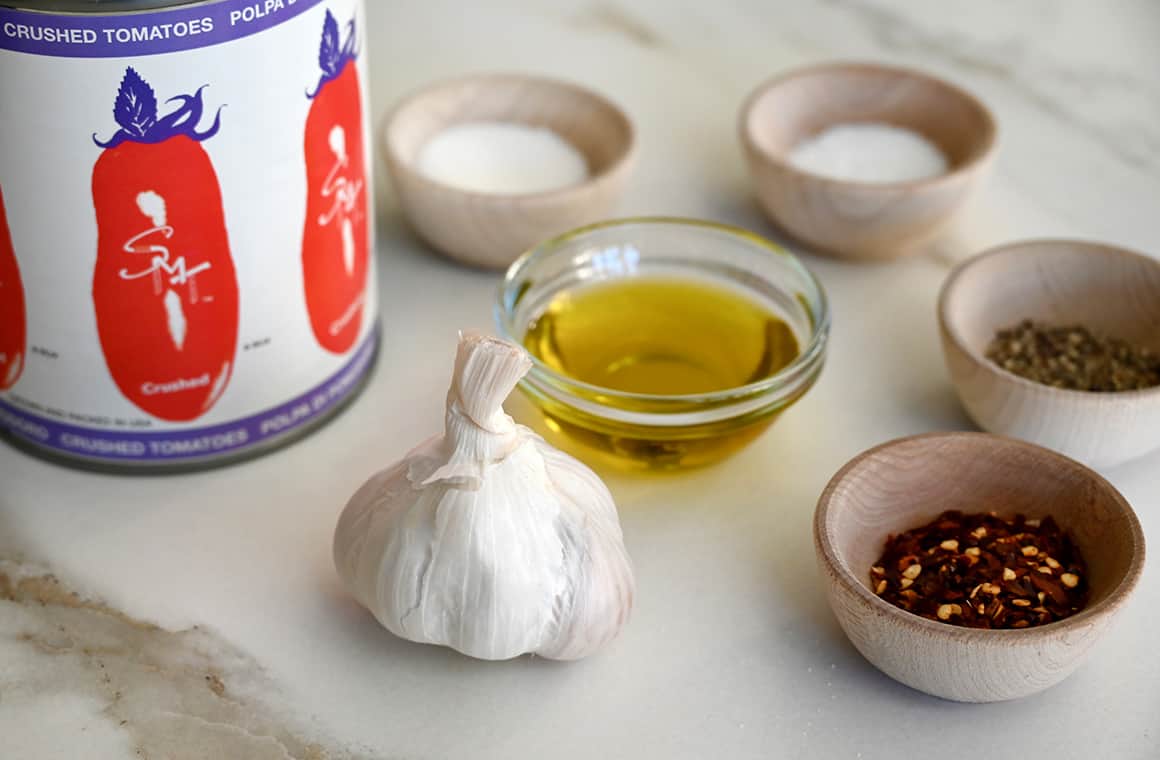 A can of Cento San Marzano crushed tomatoes next to a head of garlic and small bowls containing olive oil, crushed red pepper flakes, salt and pepper