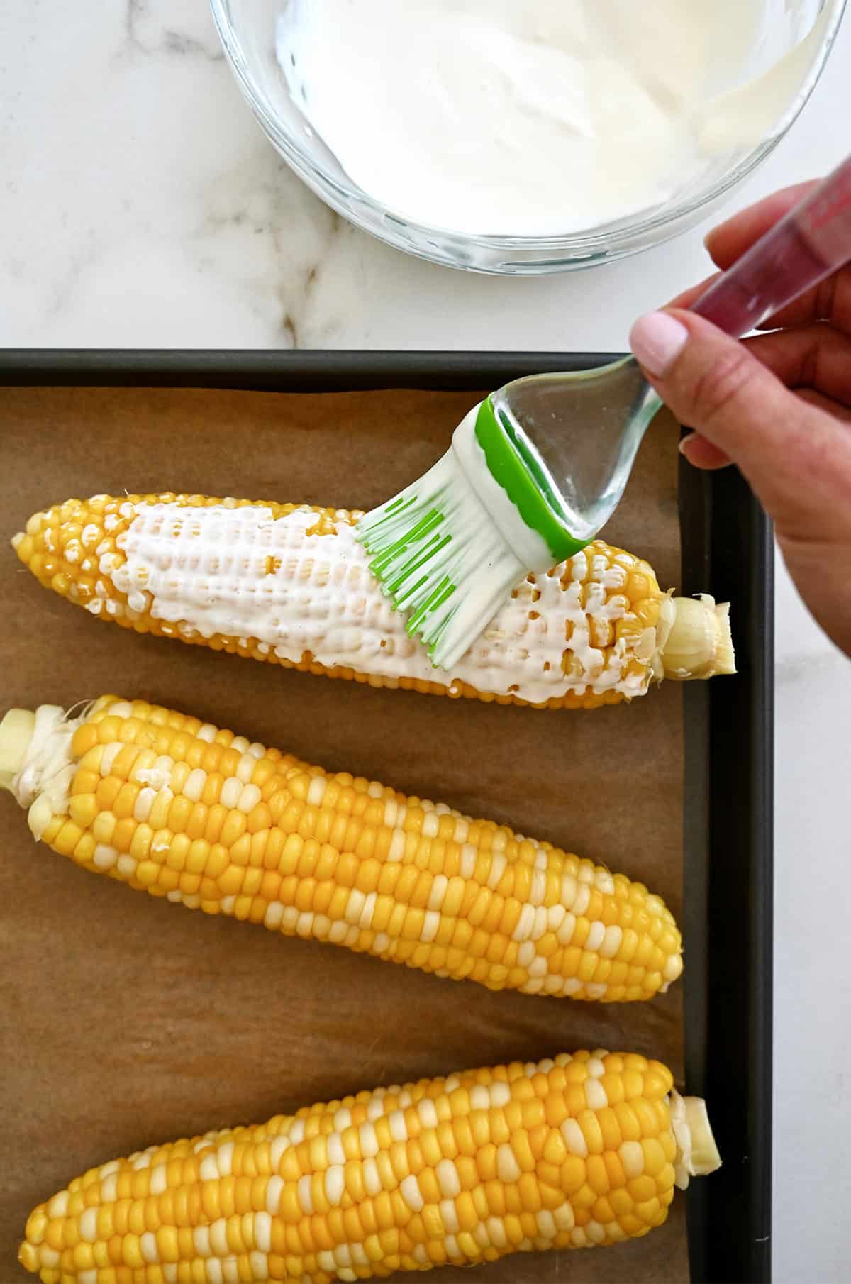 Using a silicone pastry brush to slather boiled corn cobs with a creamy sauce.