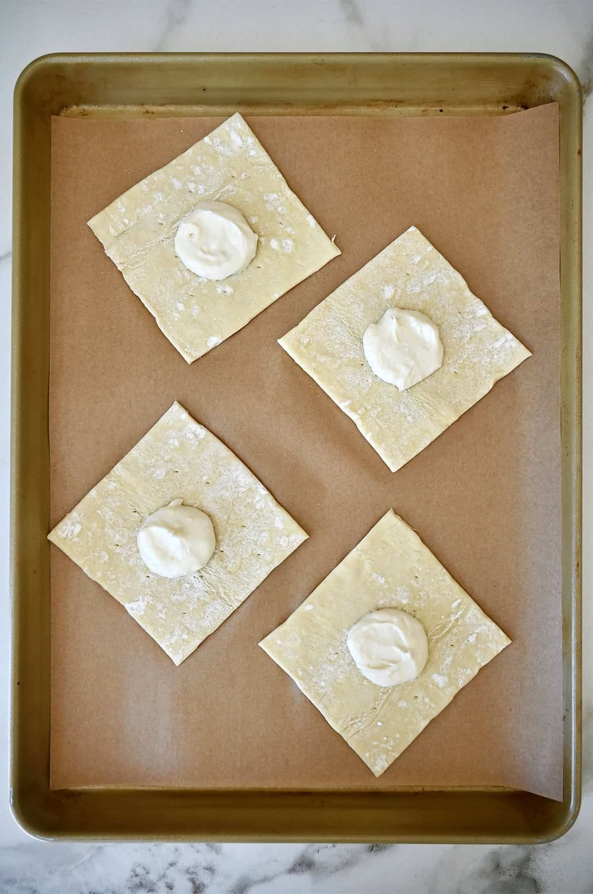 Four squares of puff pastry topped with a dollop of cream cheese on a parchment paper-lined baking sheet.