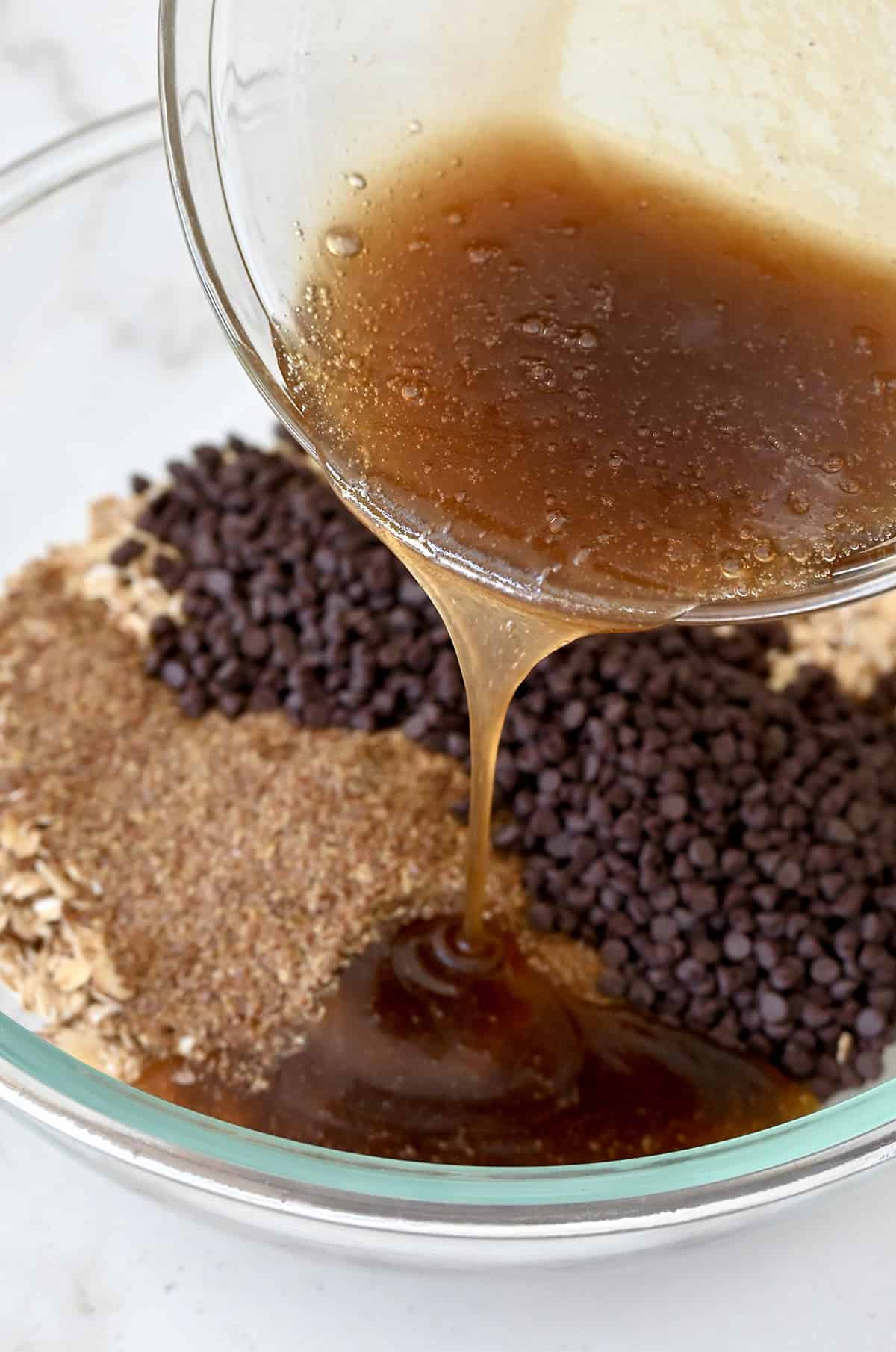 Adding wet ingredients to a mixing bowl with toasted oats, mini chocolate chips and flax meal for homemade granola bars.