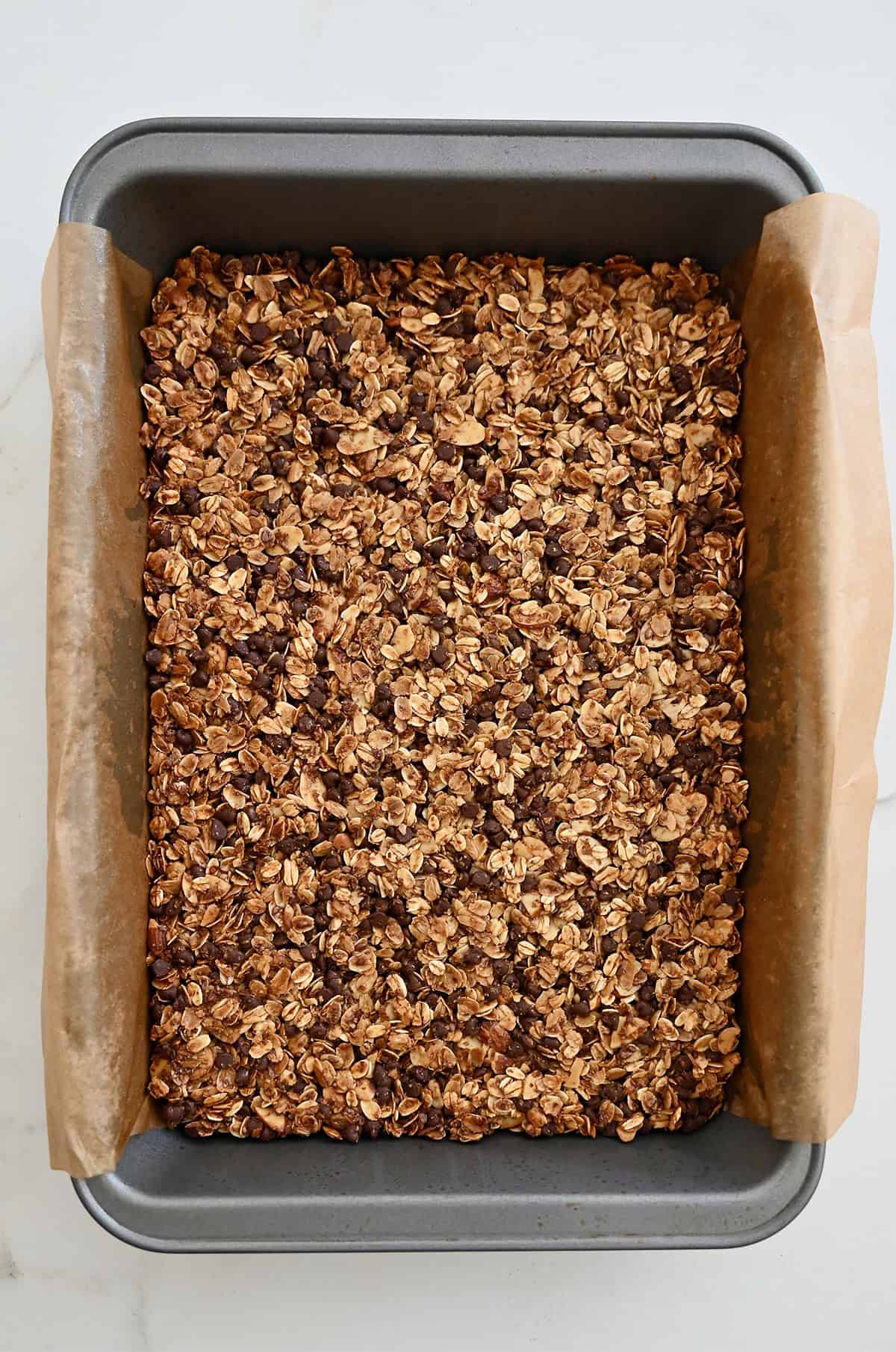 Dark golden granola bars with chocolate chips in a 9x13-inch metal pan lined with parchment paper.