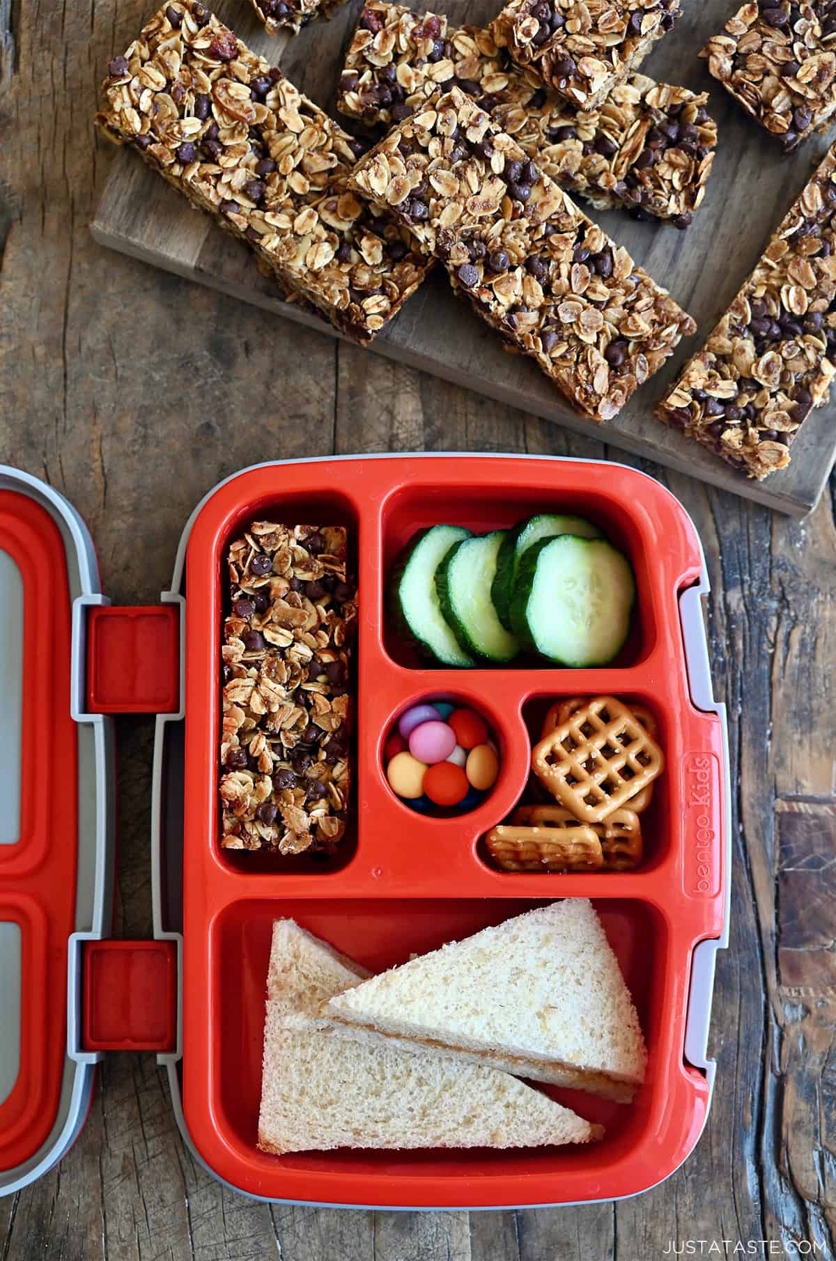Homemade granola bars on a cutting board next to a lunchbox with a granola bar tucked inside.