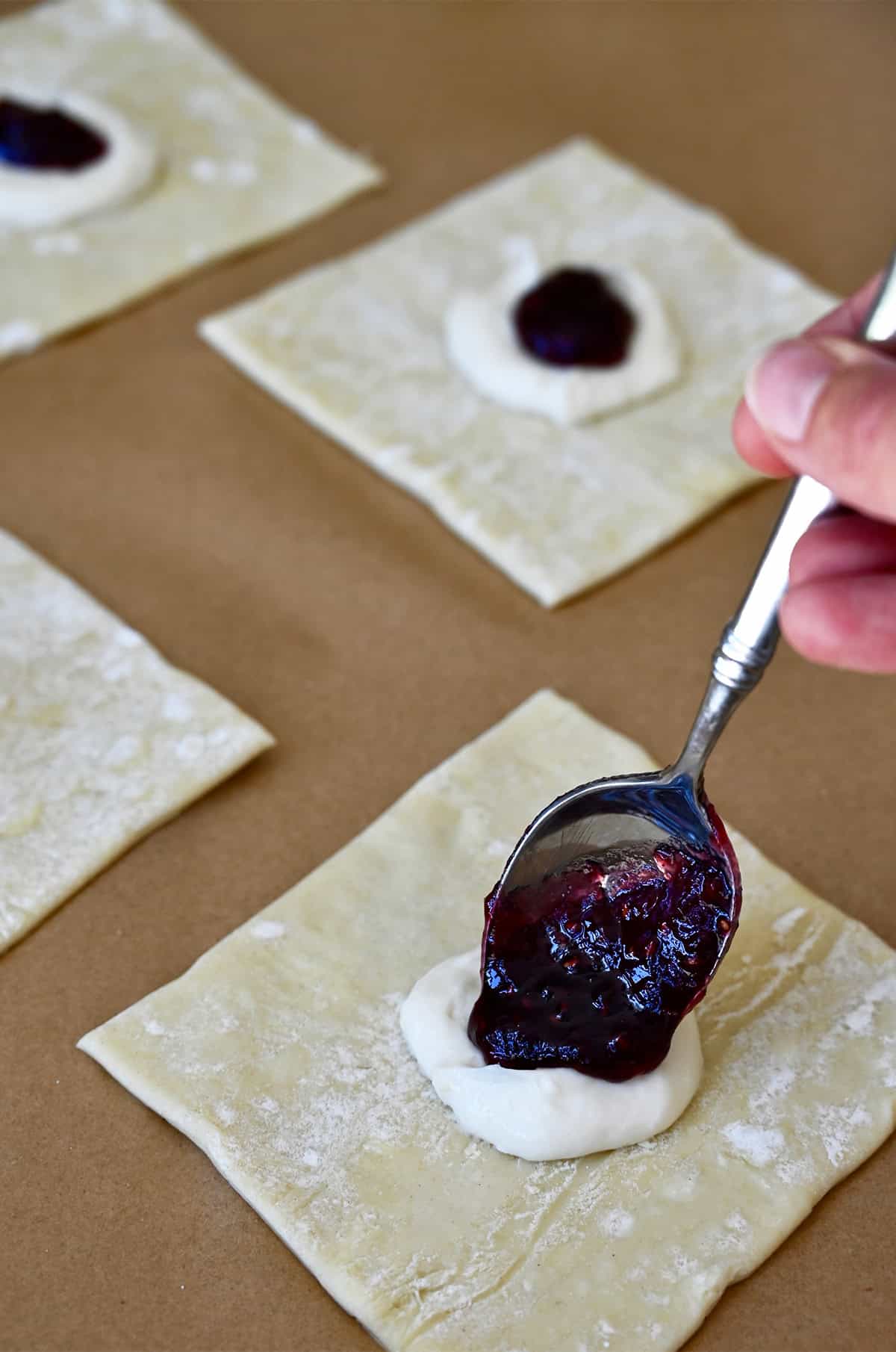 Jam being spooned atop a dollop of cream cheese on a square of puff pastry.