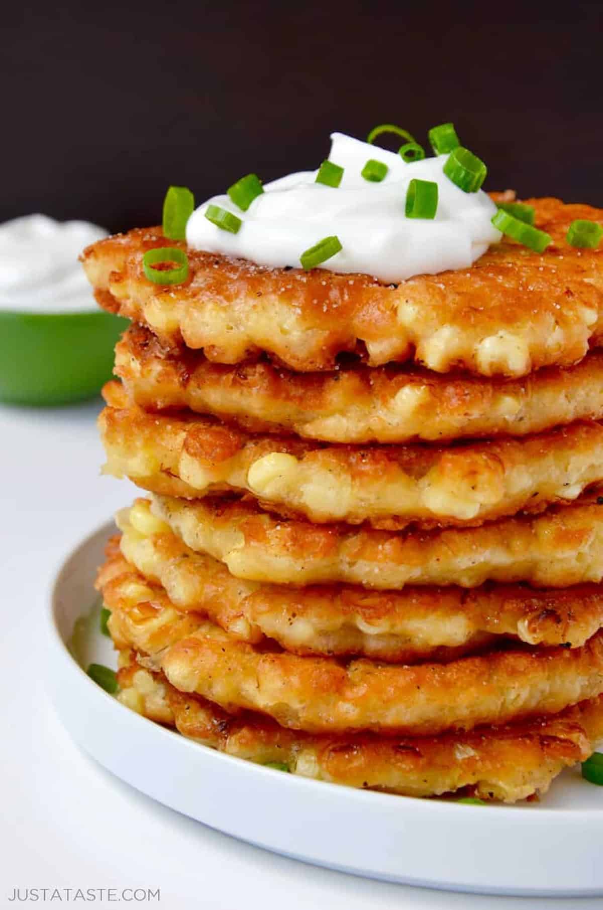 A tall stack of crispy corn fritters topped with a dollop of sour cream and chopped green onions.