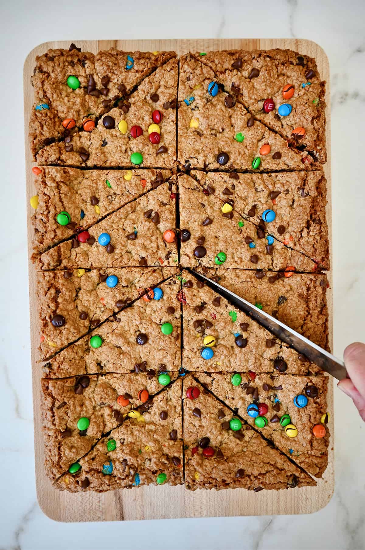 A hand holding a sharp knife cuts peanut butter oatmeal bars studded with M&Ms and chocolate chips into perfect triangles.
