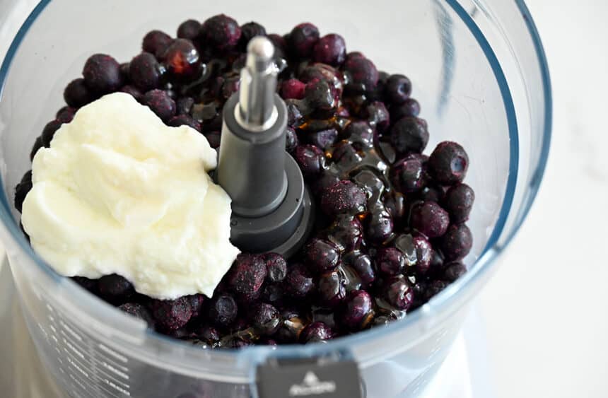 A bowl of a food processor containing frozen blueberries, honey and yogurt