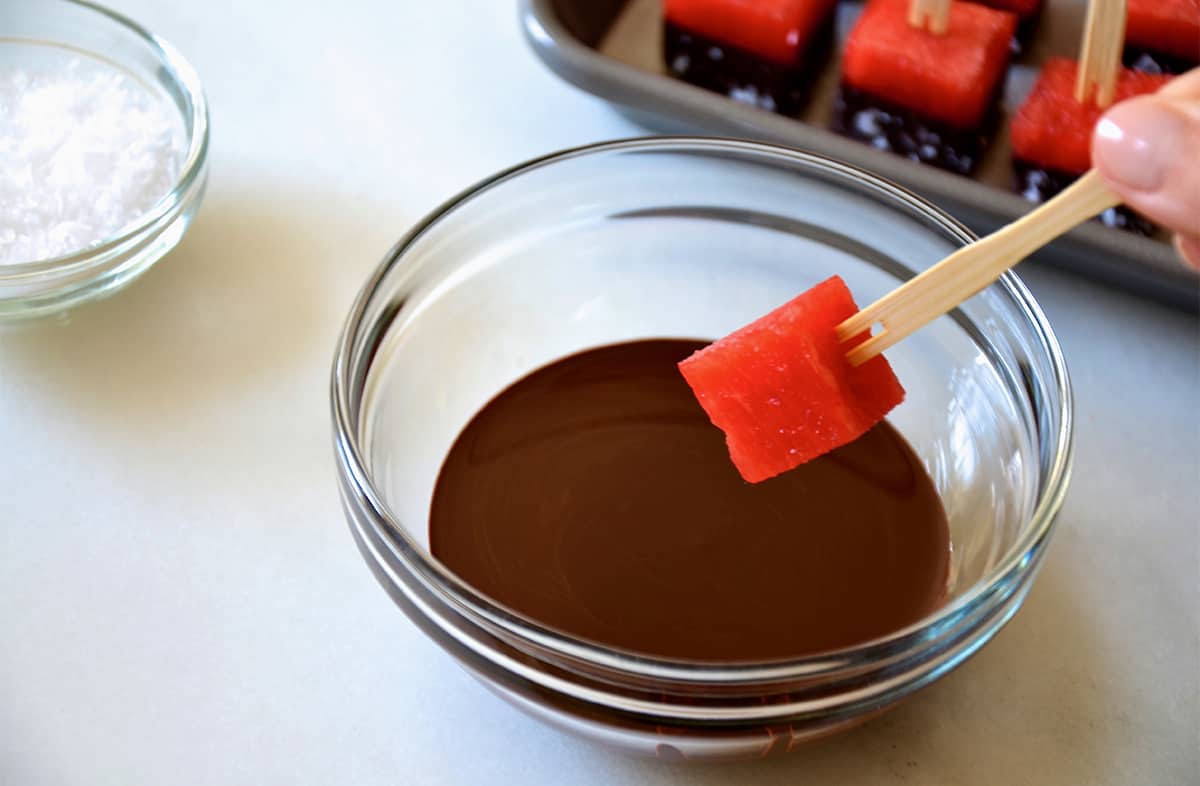 A hand holds a watermelon cube with a toothpick over a small bowl of melted chocolate.