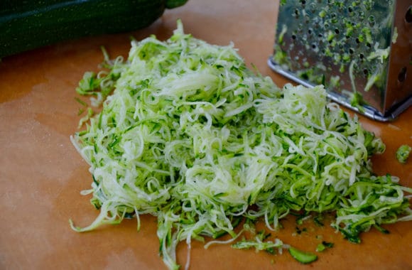 Grated zucchini pile next to box grater and zucchini