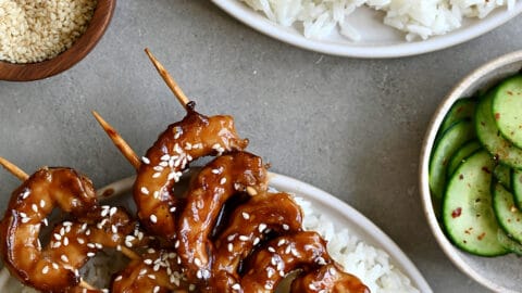 Several skewers of honey glazed shrimp garnished with sesame seeds atop coconut rice on a plate with cucumber salad.
