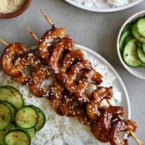 Honey garlic shrimp skewers on a plate containing steamed white rice and cucumber salad.