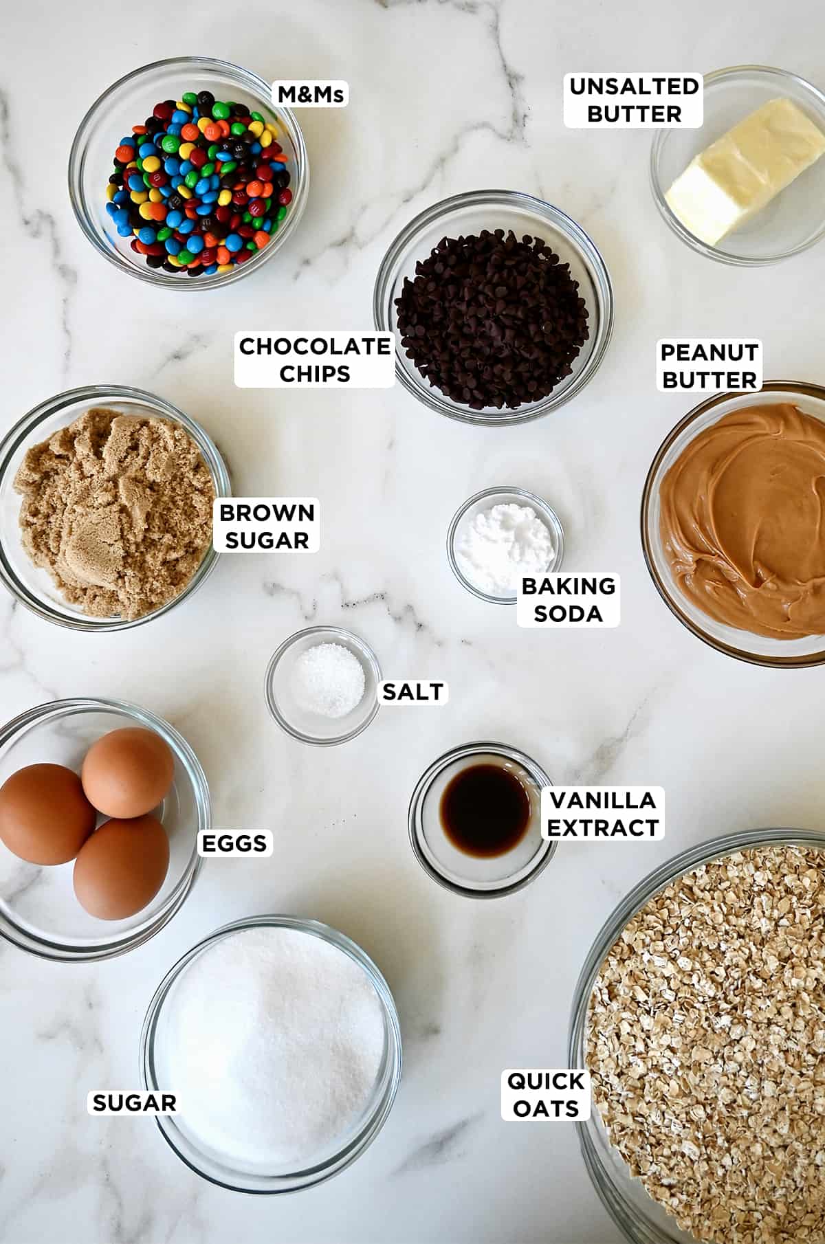 Glass bowls containing ingredients to make monster cookie bars, including M&Ms, chocolate chips, baking soda, white and brown sugars, vanilla extract, peanut butter, eggs and old-fashioned oats.