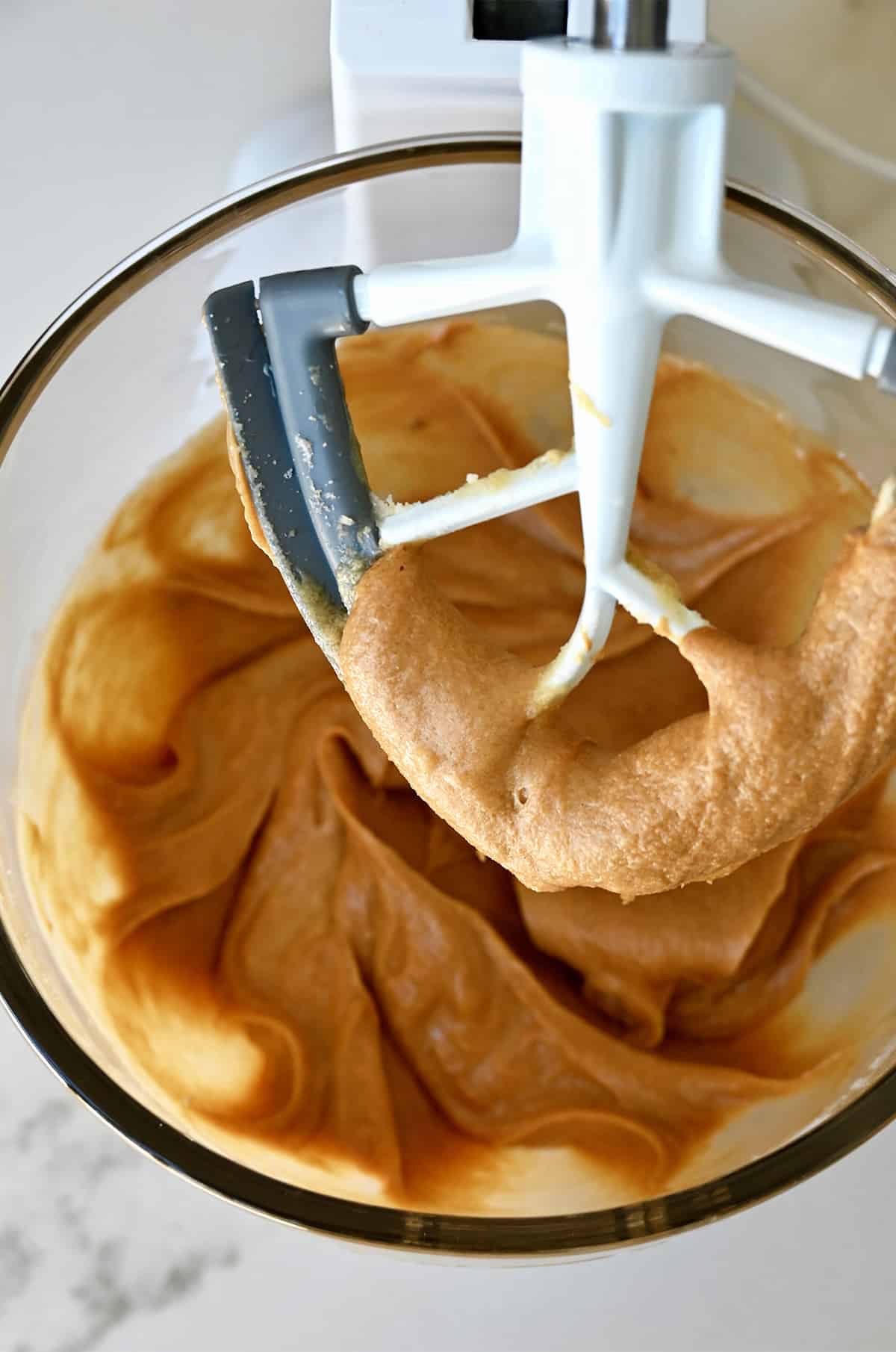 The paddle attachment of a stand mixer coated in a sugar and peanut butter mixture above a bowl containing the same mixture.