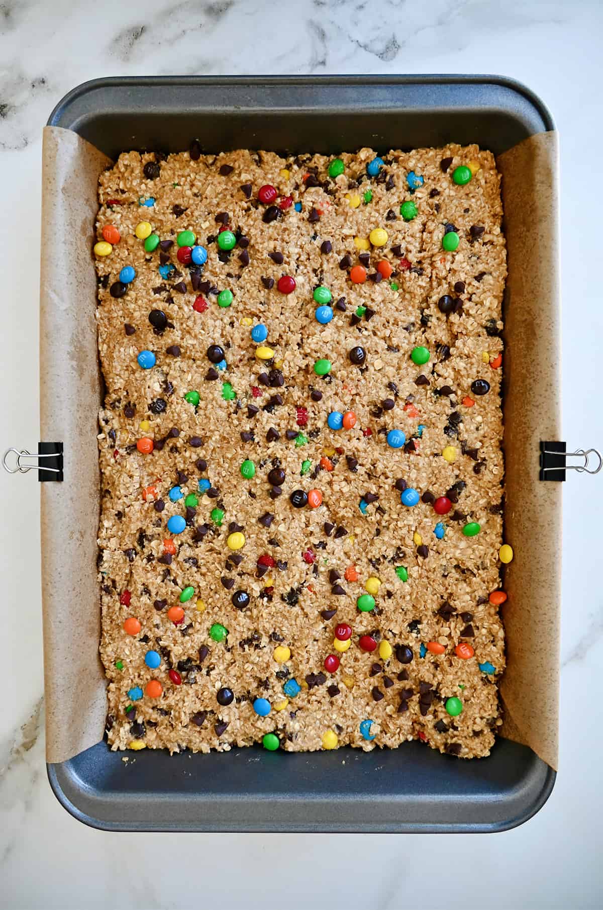 Monster cookie dough spread into an even layer in a 9x13 baking pan lined with parchment paper and secured with metal binder pins.