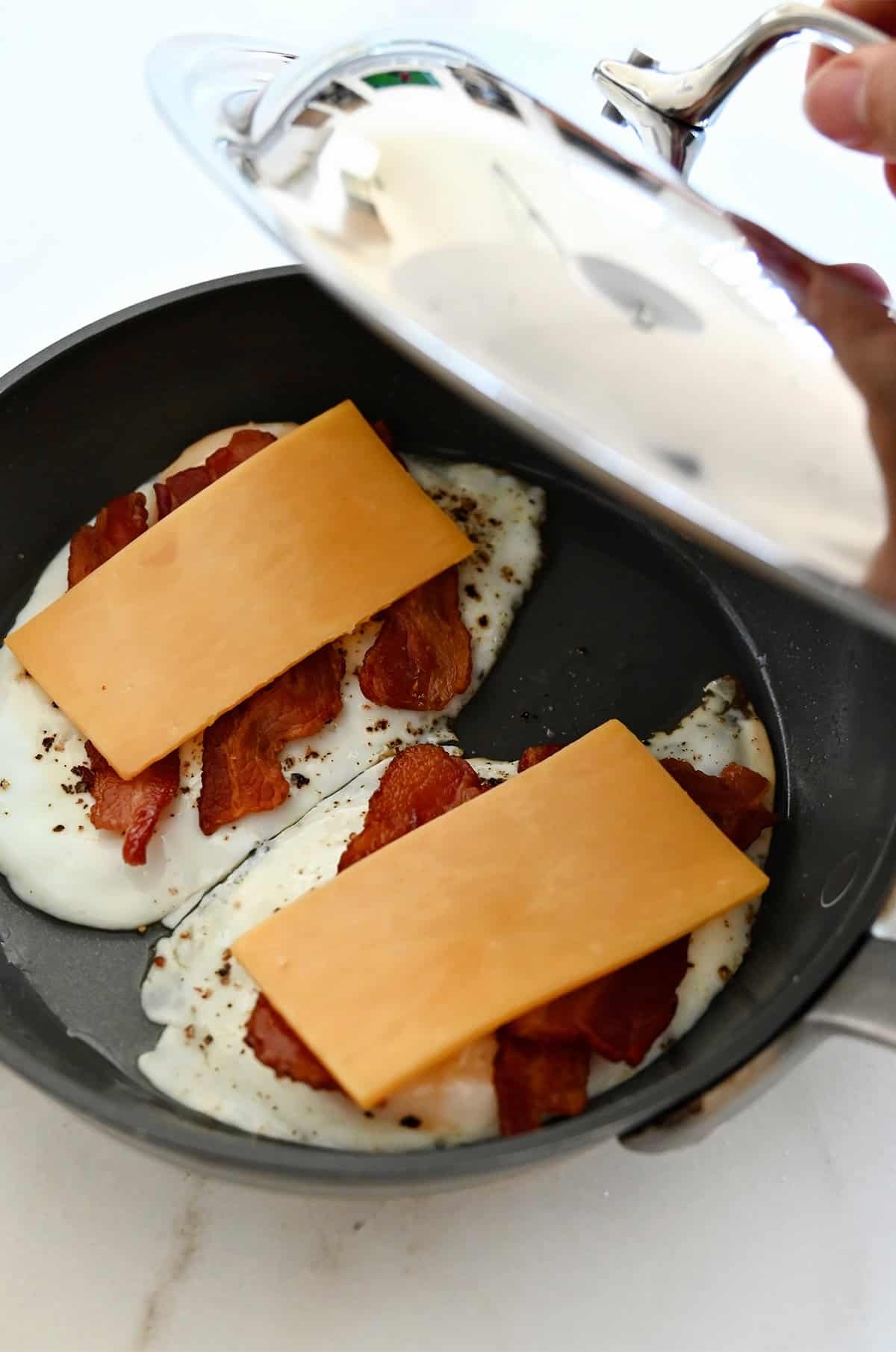 Two fried eggs topped with cooked bacon and a slice of cheddar cheese in a skillet about to be covered with a lid.