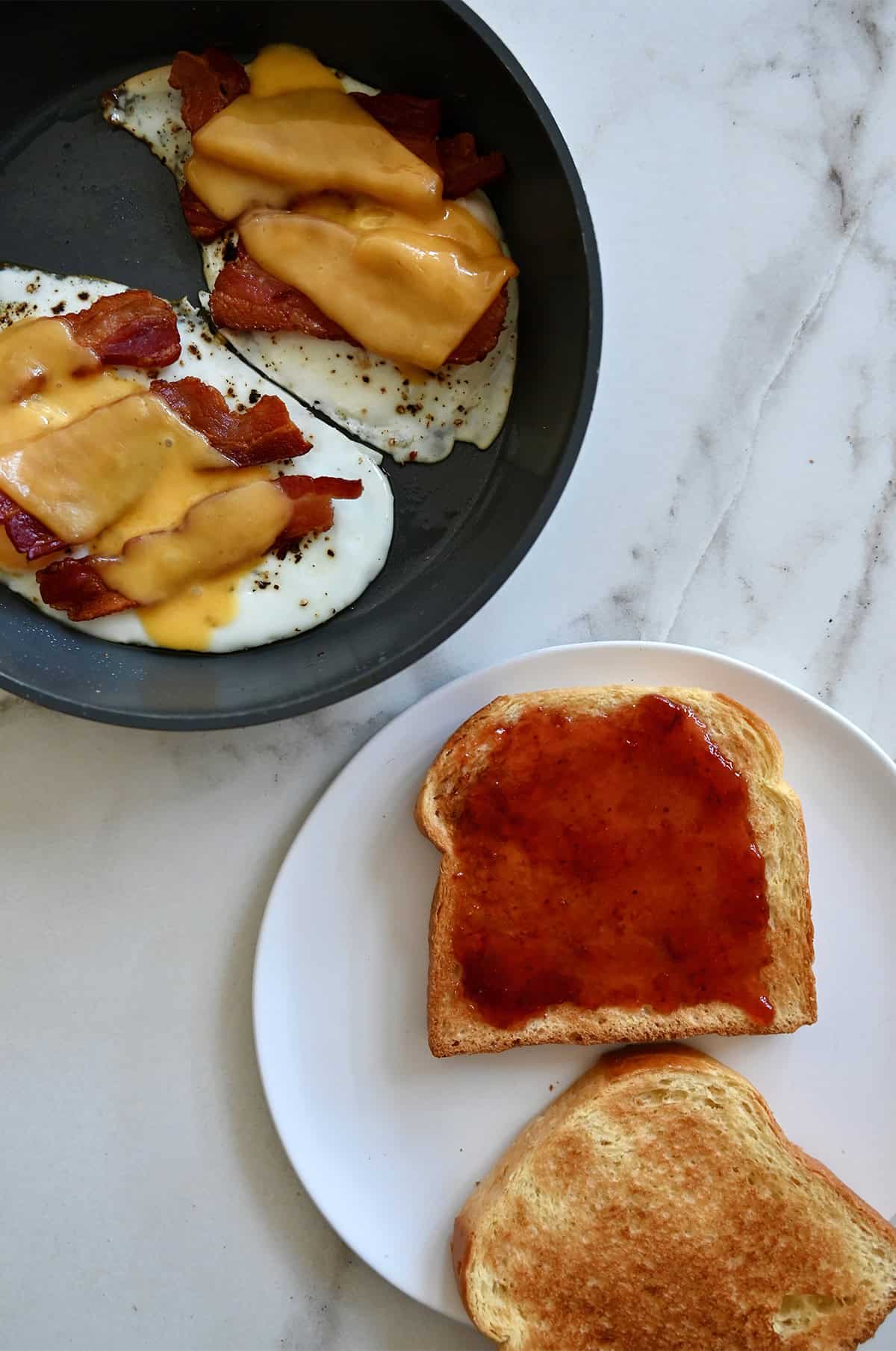 A plate with toast, one slathered with strawberry jam, next to a skillet with two fried eggs topped with crispy bacon and melted cheddar cheese.