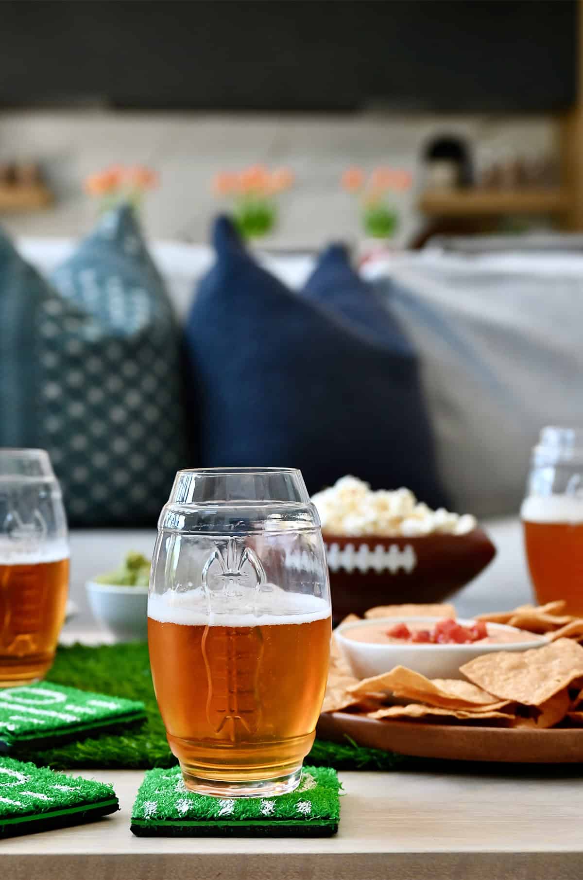 A football-shaped beer glass on an astroturf coaster surrounded by gameday snacks