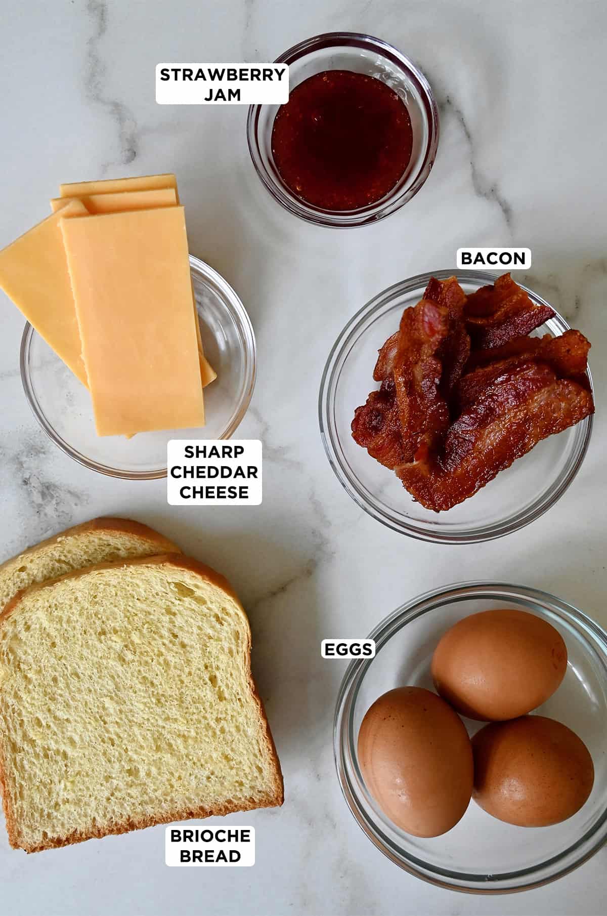 Egg sandwich ingredients in glass bowls: eggs, bacon, cheddar cheese, and strawberry jam, with two slices of bread placed beside the bowls.
