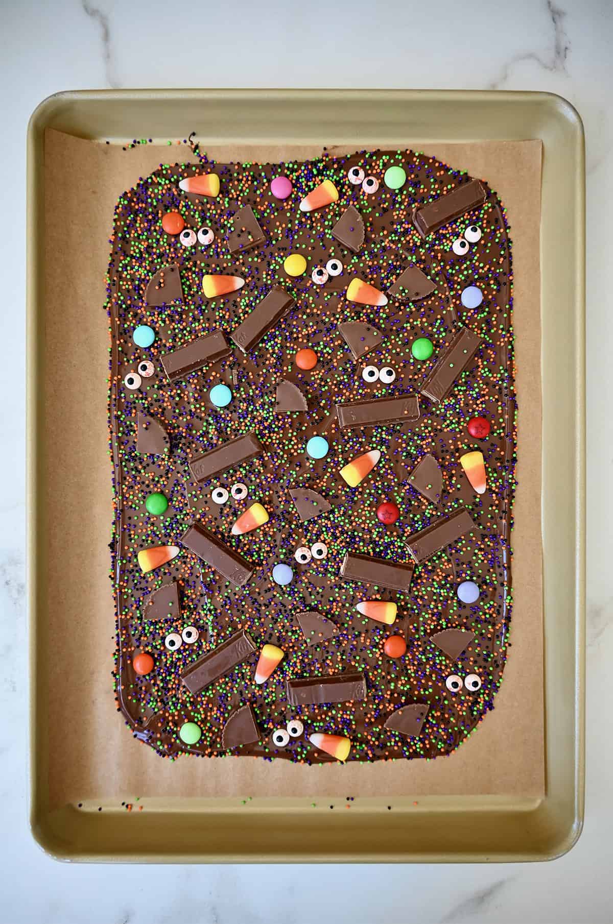 Chocolate bark topped with sprinkles, chopped candy bars, candy corn, M&Ms and bloodshot candy eyes is cooling on a parchment paper-lined baking tray.