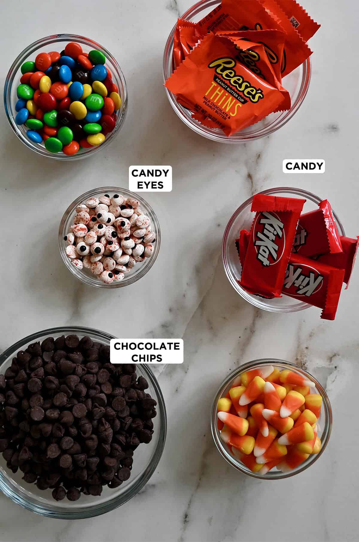 Ingredients to make Halloween chocolate candy bark, including high-quality chocolate, festive sprinkles, M&Ms, KitKat bars, candy corn, Reese's peanut butter bars and candy eyes.