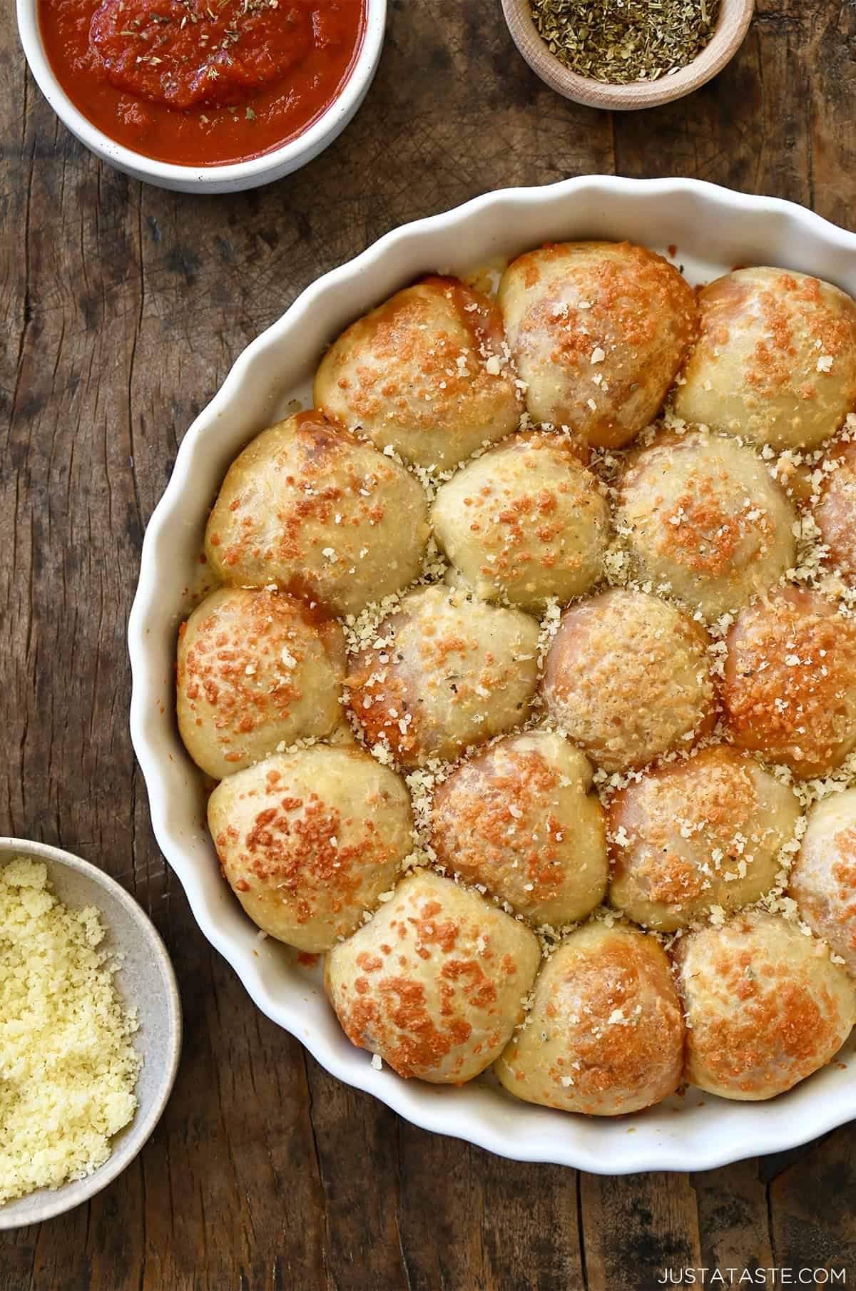 Cheesy pizza pull-apart rolls in a round pan next to small bowls containing pizza sauce and grated Parmesan.